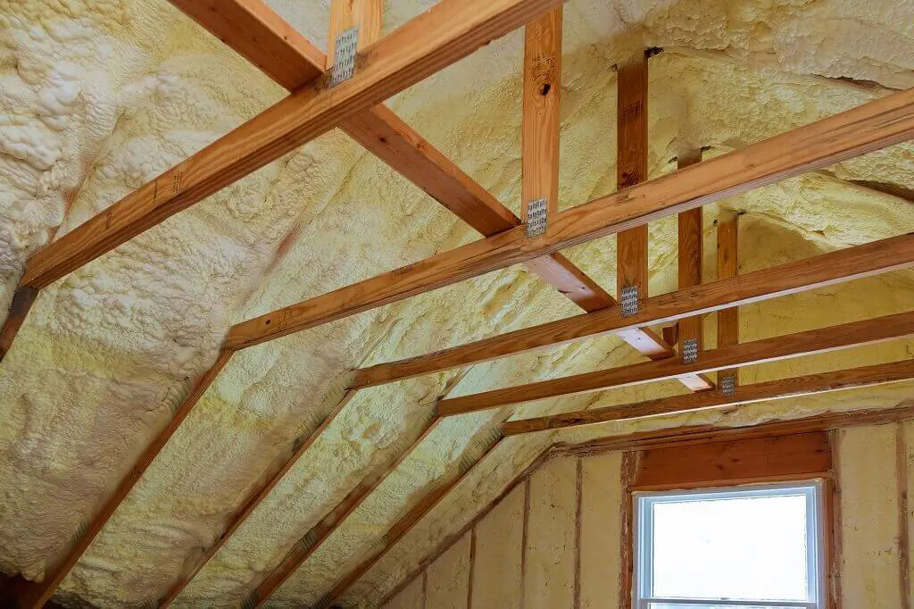 Attic roof rafters covered in sprayed yellow foam insulation with a window at the gable end