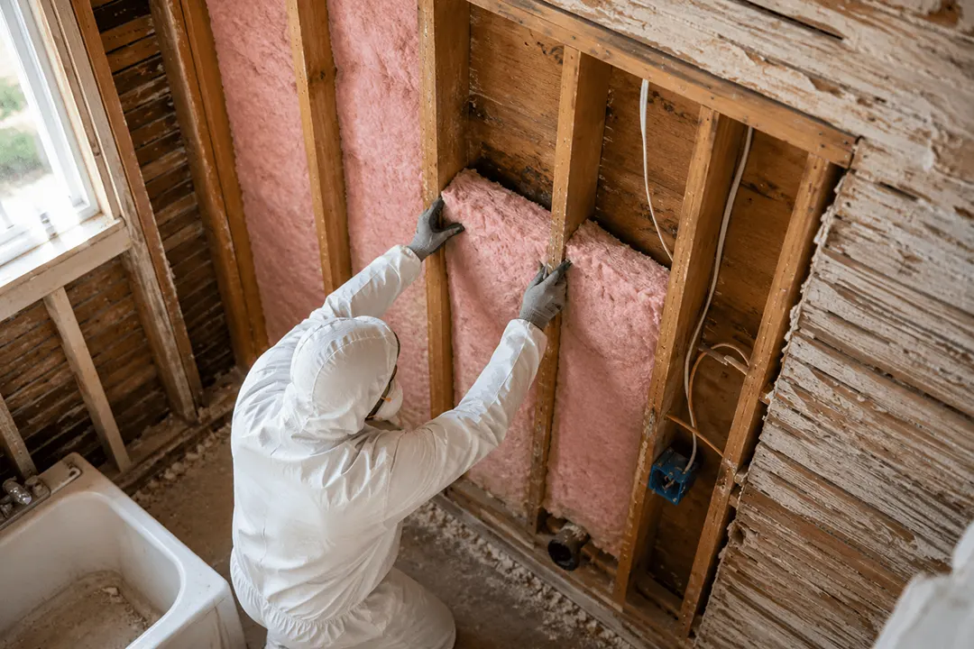 Technician in a protective suit installing pink fiberglass insulation between wall studs during a home renovation