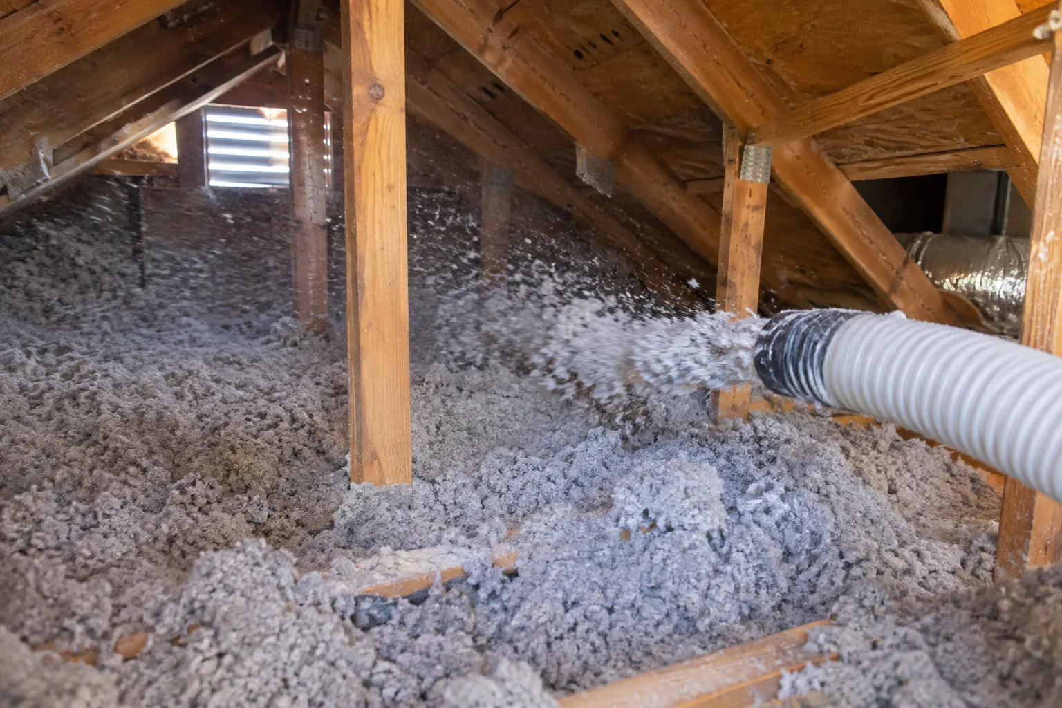 Loose-fill insulation being blown from a hose into an attic corner near ductwork, filling gaps around the framing.