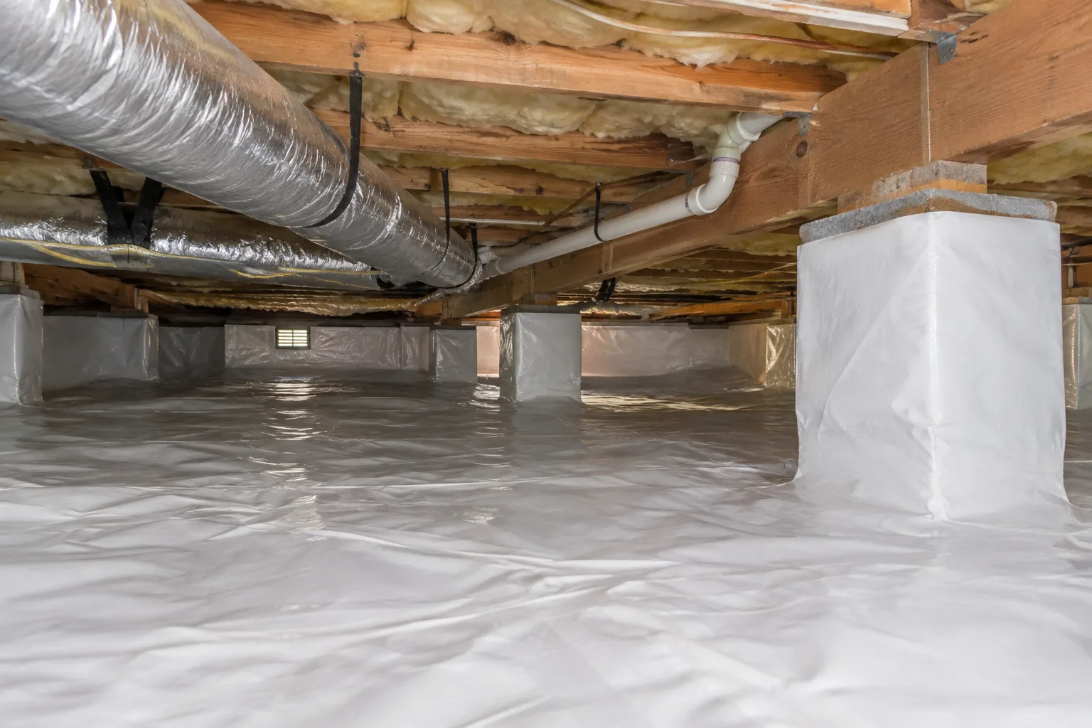 A crawl space encapsulated with a heavy-duty vapor barrier covering the floor and piers, with insulated ductwork and plumbing running overhead.