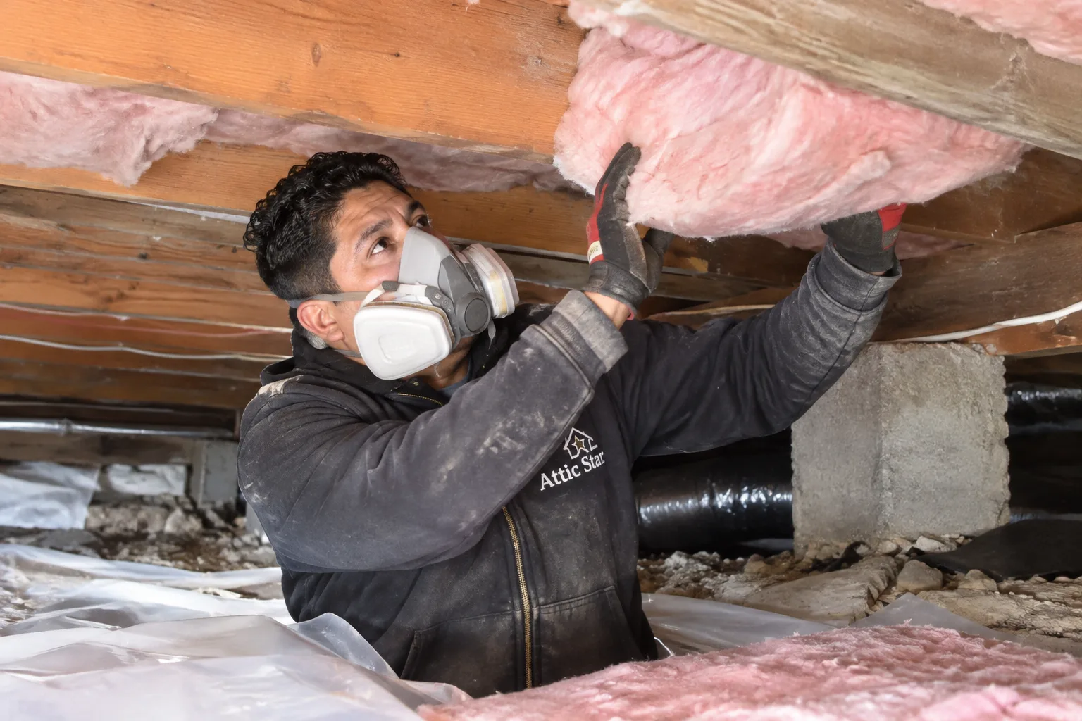 Installing new pink fiberglass batt insulation between crawl space floor joists over a sealed vapor barrier.