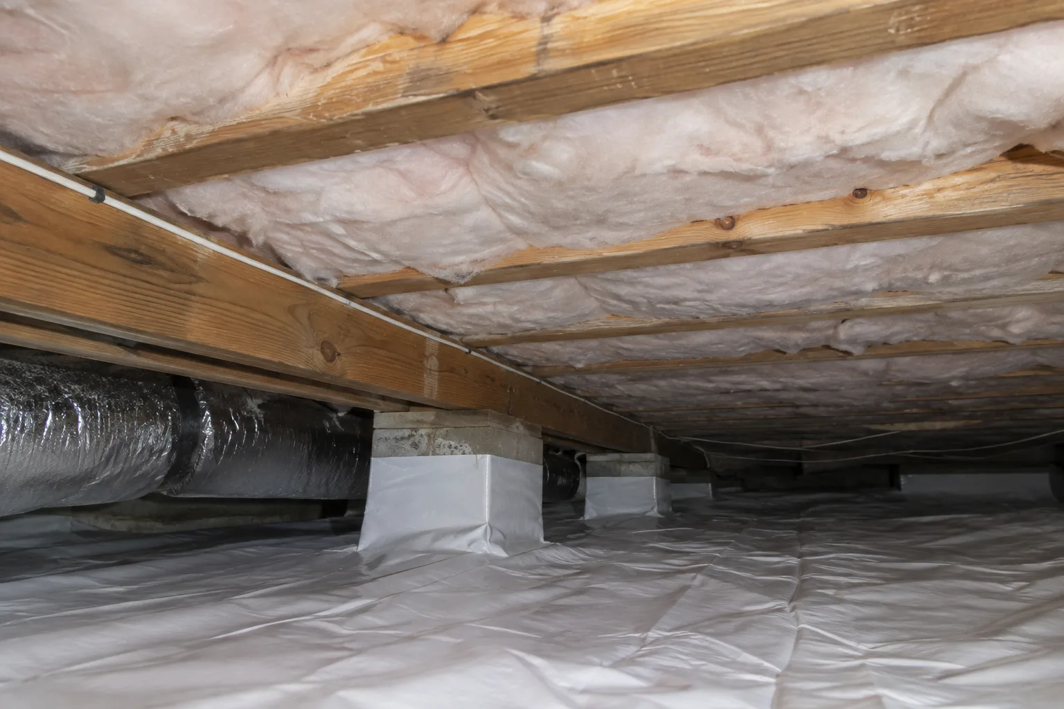 Newly installed fiberglass batt insulation spanning crawl space floor joists above a sealed vapor barrier and insulated ducting.