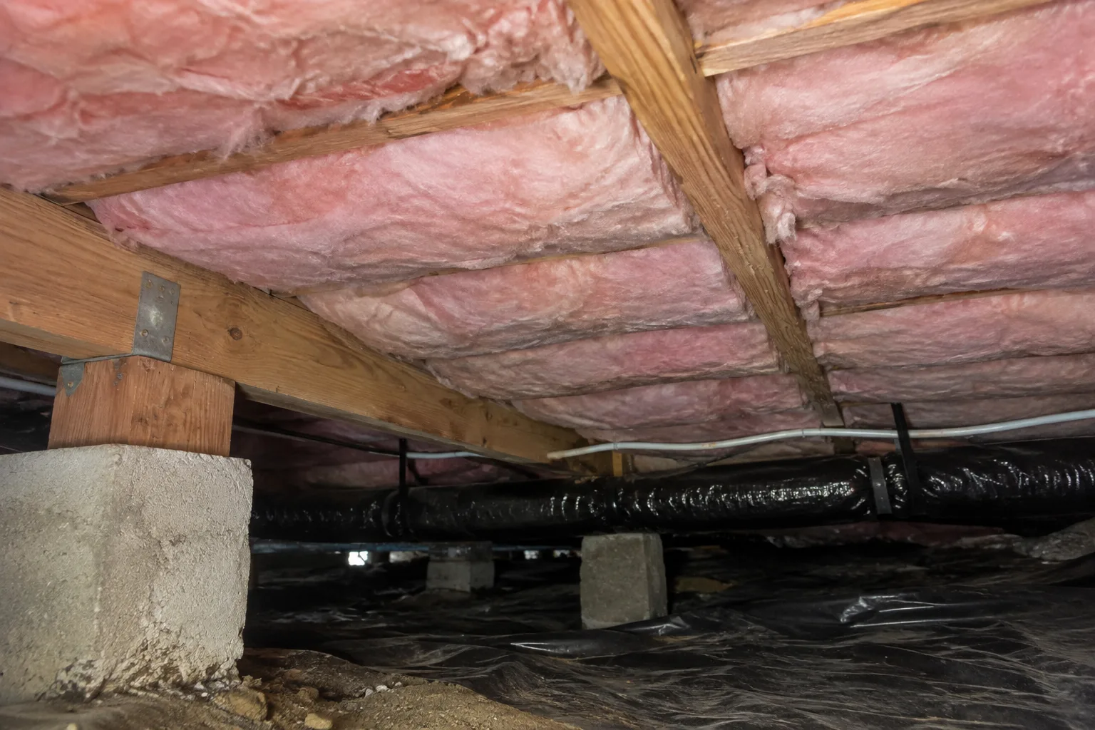 Fiberglass batt insulation installed between floor joists above a crawl space, with wrapped ductwork and a vapor barrier visible below.
