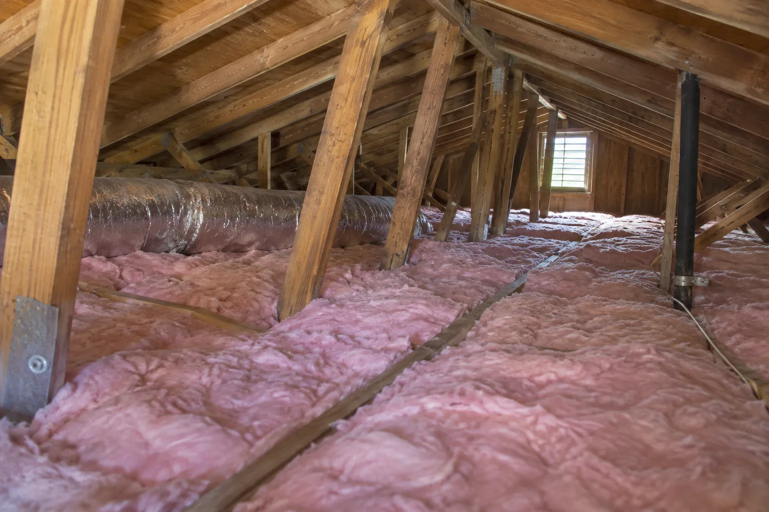 Freshly installed pink fiberglass batt insulation filling attic floor joist bays around exposed wooden framing and ductwork.
